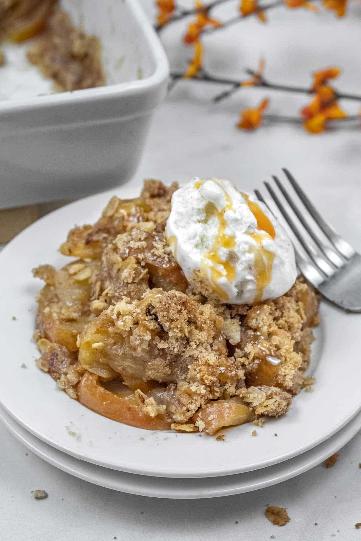 A serving of apple crisp topped with ice cream and caramel sauce sits on a white plate next to a fork.