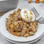 A serving of apple crisp topped with ice cream and caramel sauce sits on a white plate with a fork beside it.