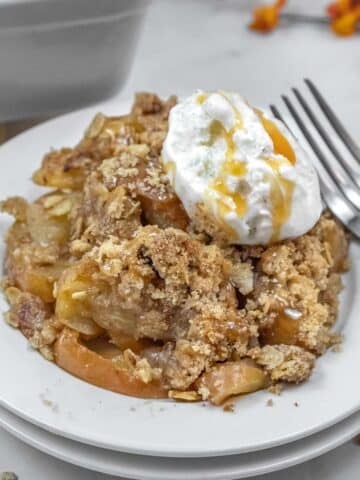 A serving of apple crisp topped with ice cream and caramel sauce sits on a white plate with a fork beside it.