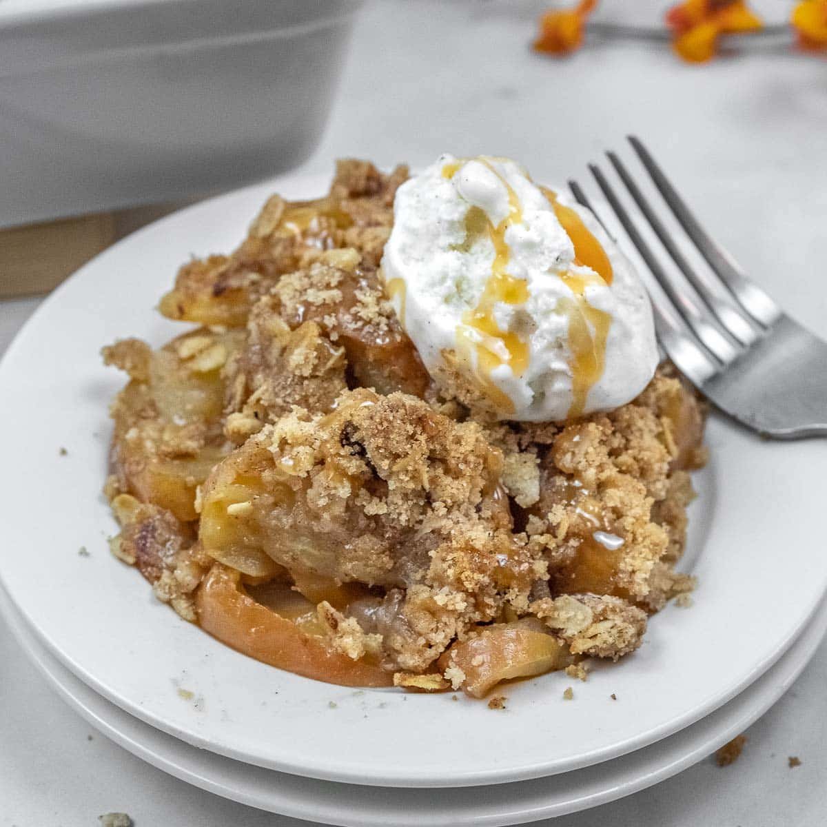 A serving of apple crisp topped with ice cream and caramel sauce sits on a white plate with a fork beside it.