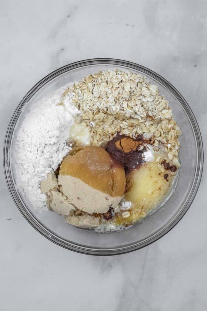A glass bowl containing oats, brown sugar, white sugar, flour, baking powder and salt, and cinnamon with melted butter poured over on a marble surface.