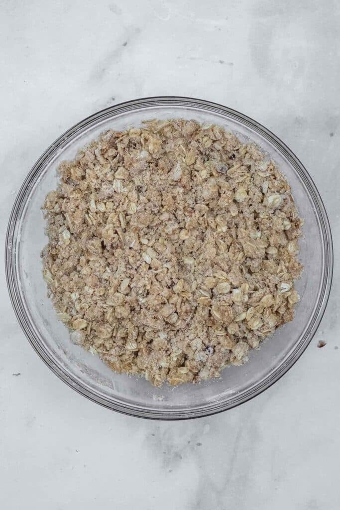 A clear glass bowl filled with an oat and brown sugar crumble mixture sits on a light gray marble surface.