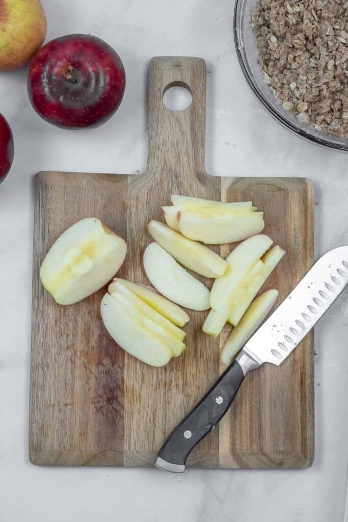 Sliced apples and a knife on a wooden cutting board, with whole apples and a bowl of crumb topping nearby.