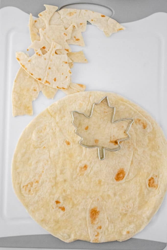 A large flour tortilla with a maple leaf-shaped cookie cutter sits on a white cutting board.