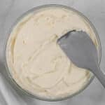 A glass mixing bowl filled with cream cheese frosting with a gray spatula inserted.