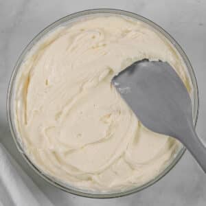A glass mixing bowl filled with cream cheese frosting with a gray spatula inserted.