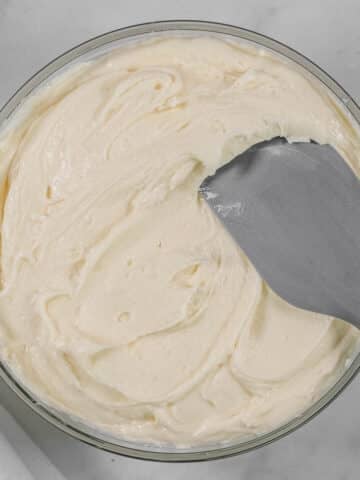 A glass mixing bowl filled with cream cheese frosting with a gray spatula inserted.