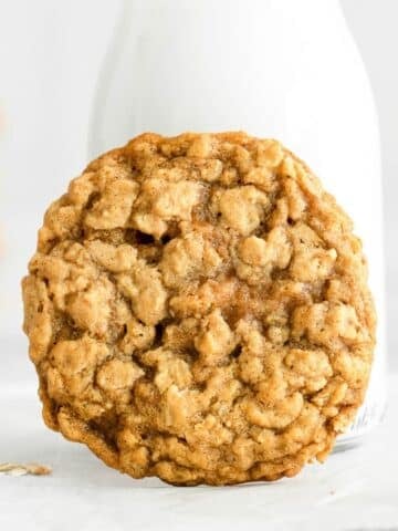 A single oatmeal cookie rests in front of a glass bottle of milk on a white surface.