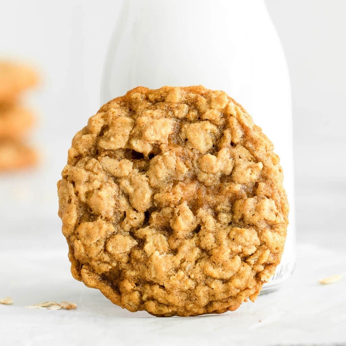A single oatmeal cookie rests in front of a glass bottle of milk on a white surface.