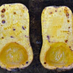 Two halves of a roasted butternut squash are placed side by side on a dark baking sheet, showing browned edges and slightly caramelized surfaces.