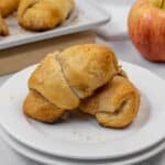 Two golden-brown apple pie crescent rolls on a white plate, with more crescent rolls on a tray and a red apple in the background.