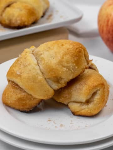 Two golden-brown apple pie crescent rolls on a white plate, with more crescent rolls on a tray and a red apple in the background.