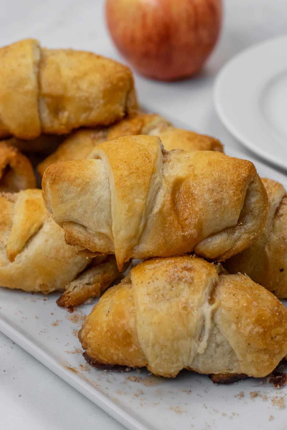 A stack of apple pie crescent rolls on a white plate, with a red apple and a white dish in the background.