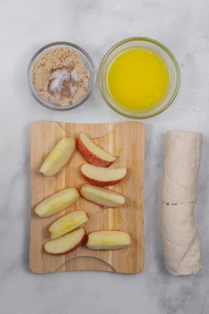 Sliced apples on a wooden board, a roll of crescent roll dough, a bowl of melted butter, and a bowl of brown sugar mixed with white sugar on a white surface.