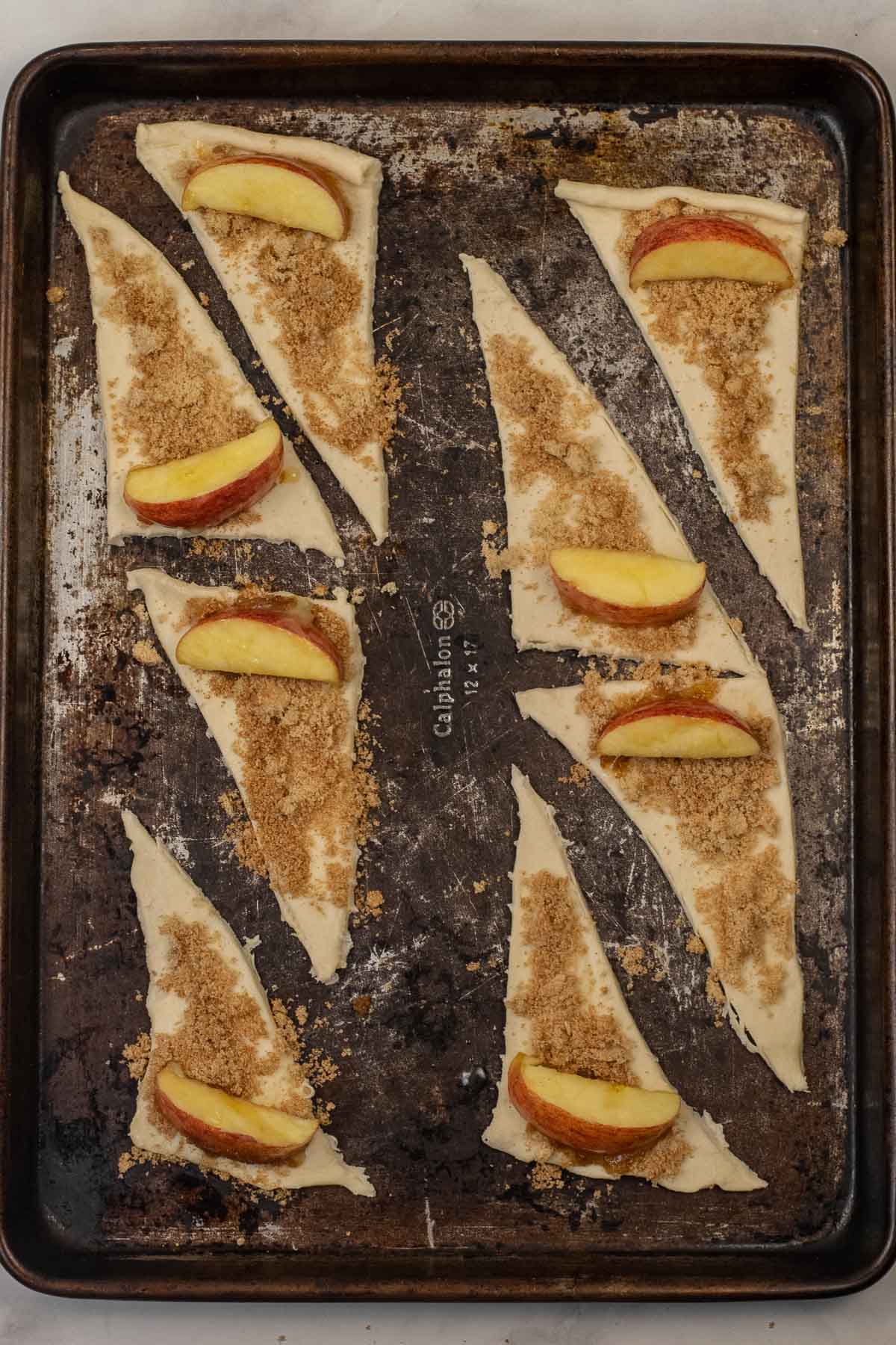 Eight unbaked crescent roll triangles on a baking sheet, each topped with brown sugar and a slice of apple.