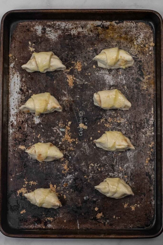 Eight unbaked crescent rolls evenly spaced on a worn, dark baking sheet, ready to be baked.