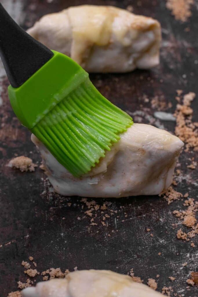 A green silicone brush applies butter to a piece of dough on a dark baking sheet, with scattered brown sugar around.
