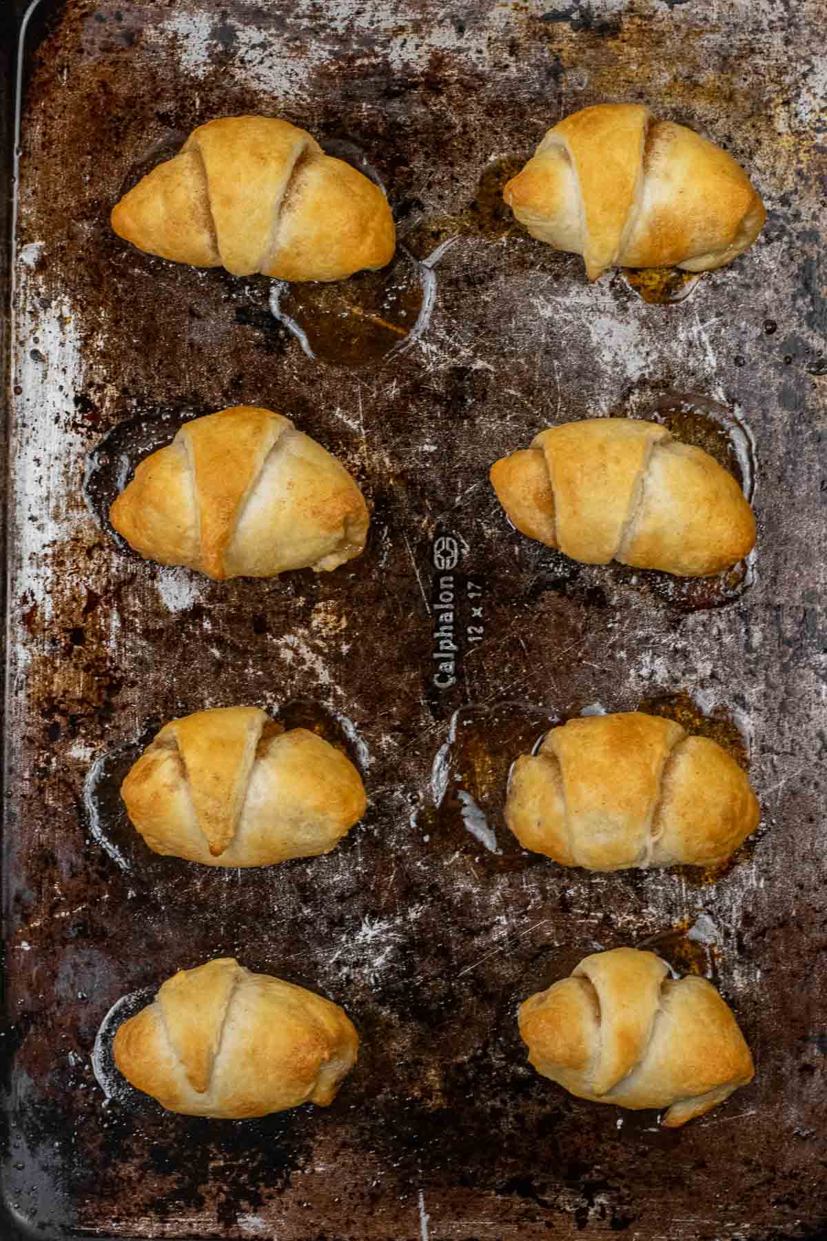 Eight golden-brown apple pie crescent rolls are baked and arranged in two rows on a worn, dark baking sheet.