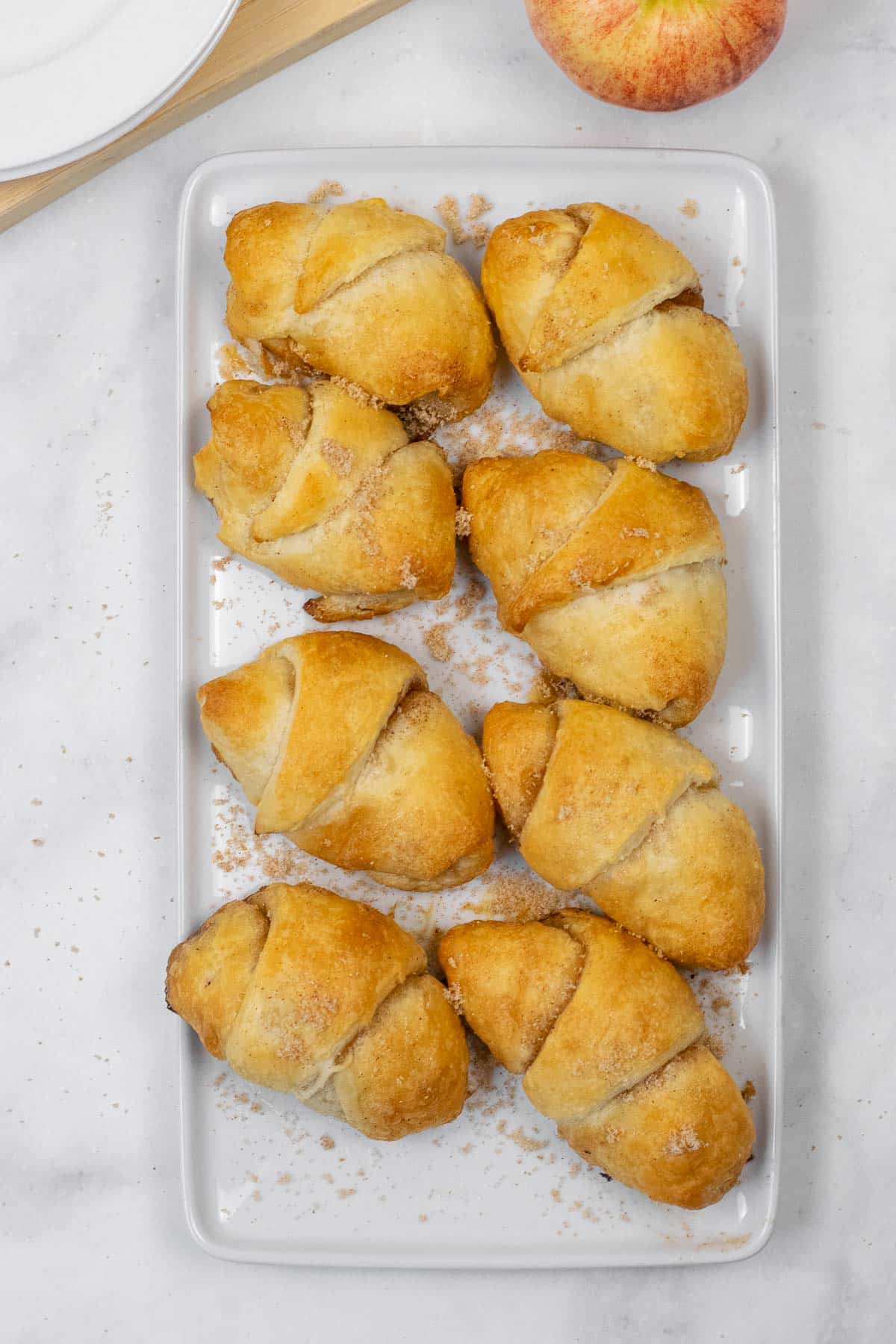 Eight golden-brown apple pie crescent rolls are arranged on a rectangular white plate.