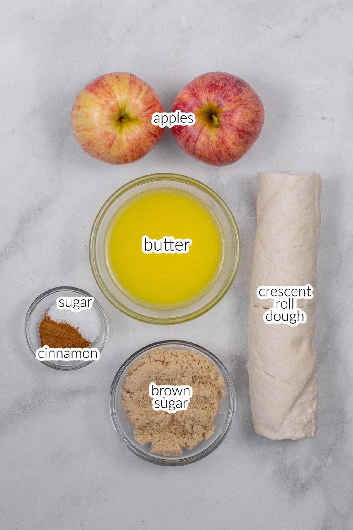 Top-down view ingredients for apple pie crescent rolls including two apples, a bowl of melted butter, a roll of crescent dough, a bowl of brown sugar, a bowl of white sugar, and a small bowl of cinnamon on a gray surface.