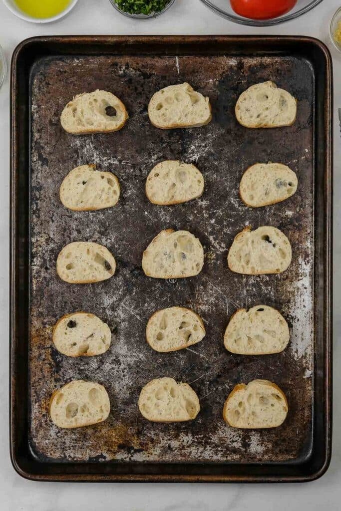 A baking sheet with fifteen slices of baguette arranged in rows, ready to be toasted.