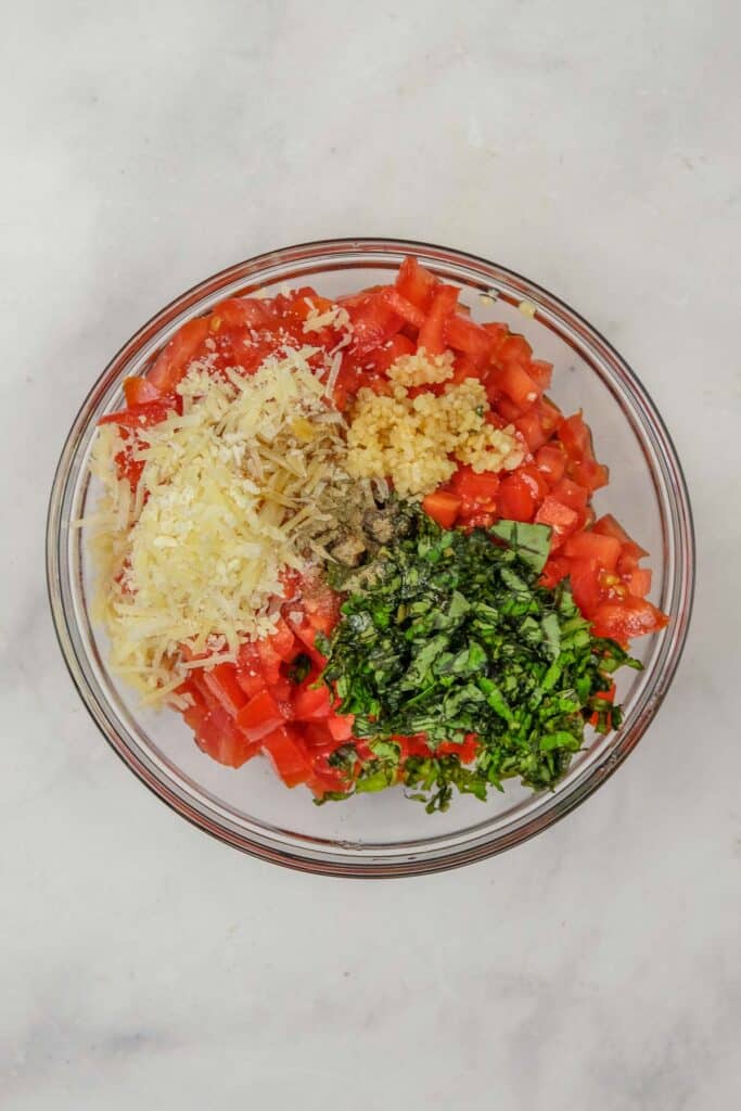 A glass bowl containing chopped tomatoes, shredded cheese, minced garlic, chopped basil, salt, and ground black pepper on a light surface.