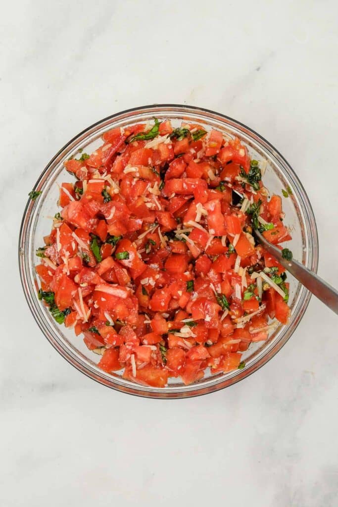 A glass bowl filled with mixed chopped tomatoes, shredded cheese, minced garlic, chopped basil, salt, and ground black pepper.