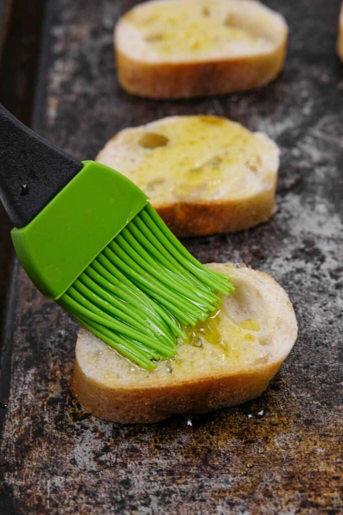 A green silicone brush spreads olive oil on a slice of baguette bread placed on a baking sheet.