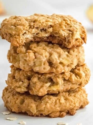 A stack of four apple oatmeal cookies, with the top cookie showing a bite taken out of it, on a white surface.
