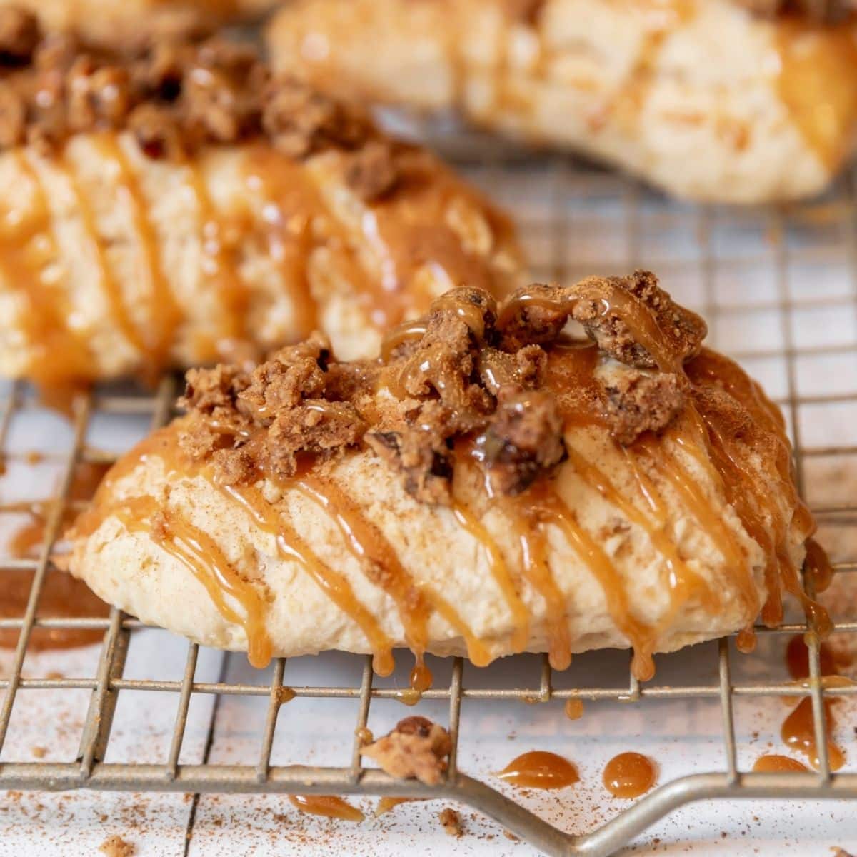 A scone topped with crumbled topping and drizzled caramel glaze sits on a wire cooling rack.