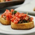 Two slices of toasted bread topped with a mixture of diced tomatoes, basil, garlic, and olive oil are served on a white plate.