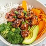 A bowl with white rice, broccoli florets, sliced avocado, carrot ribbons, and glazed teriyaki salmon cubes garnished with sesame seeds and chopped green onions.