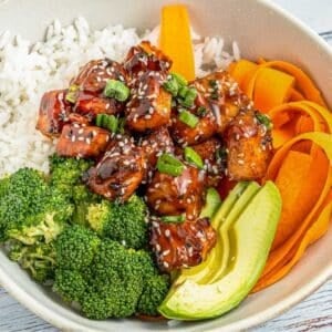A bowl with white rice, broccoli florets, sliced avocado, carrot ribbons, and glazed teriyaki salmon cubes garnished with sesame seeds and chopped green onions.