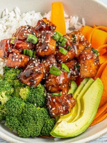 A bowl with white rice, broccoli florets, sliced avocado, carrot ribbons, and glazed teriyaki salmon cubes garnished with sesame seeds and chopped green onions.