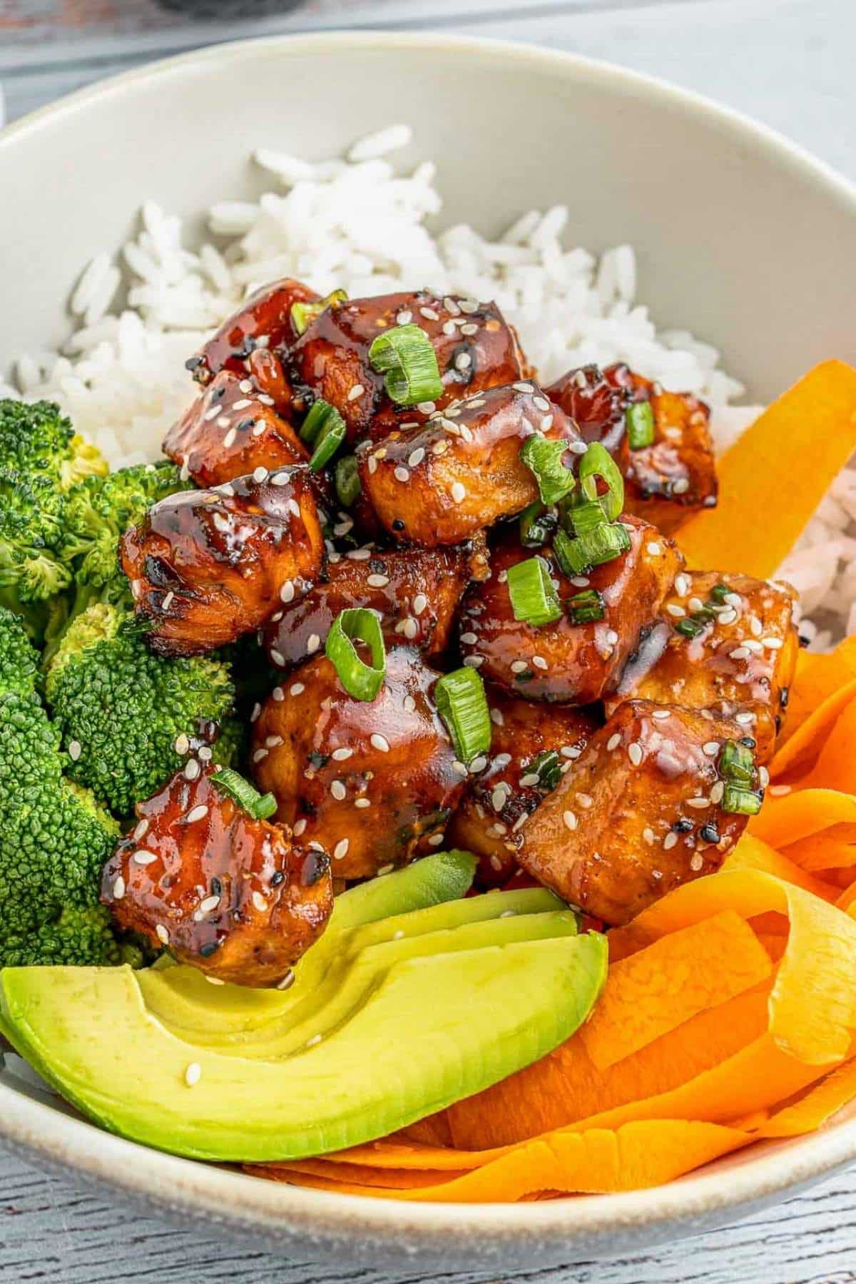 A bowl with white rice, glazed tofu cubes topped with green onions and sesame seeds, sliced avocado, broccoli, and carrot ribbons.
