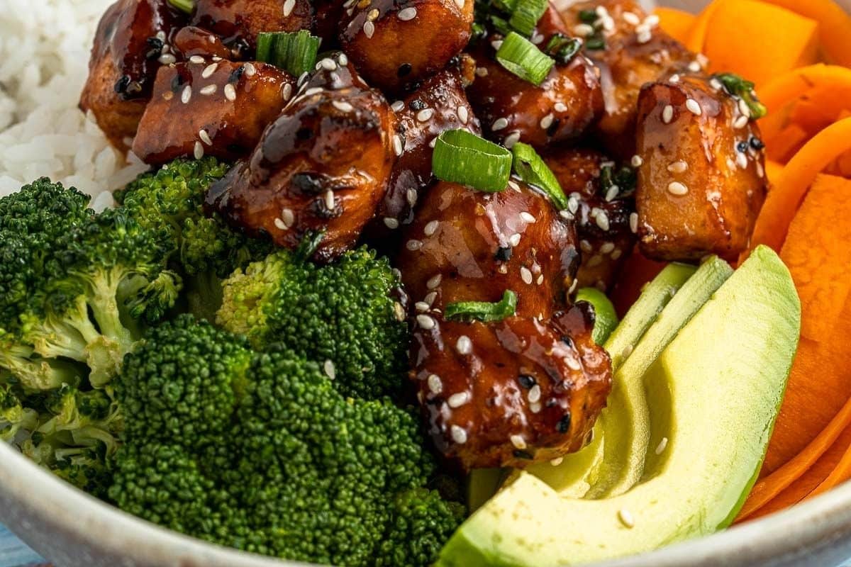 A bowl with glazed tofu, steamed broccoli, sliced avocado, carrots, white rice, and a sprinkle of sesame seeds and green onions.