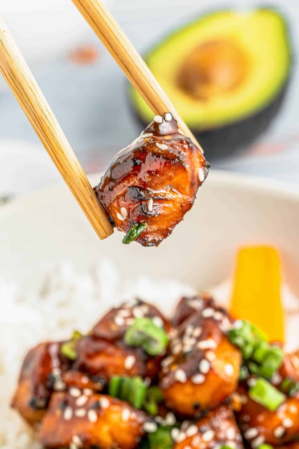 Chopsticks hold a glazed salmon cube topped with sesame seeds and green onion, with a bowl of rice and sliced avocado in the background.