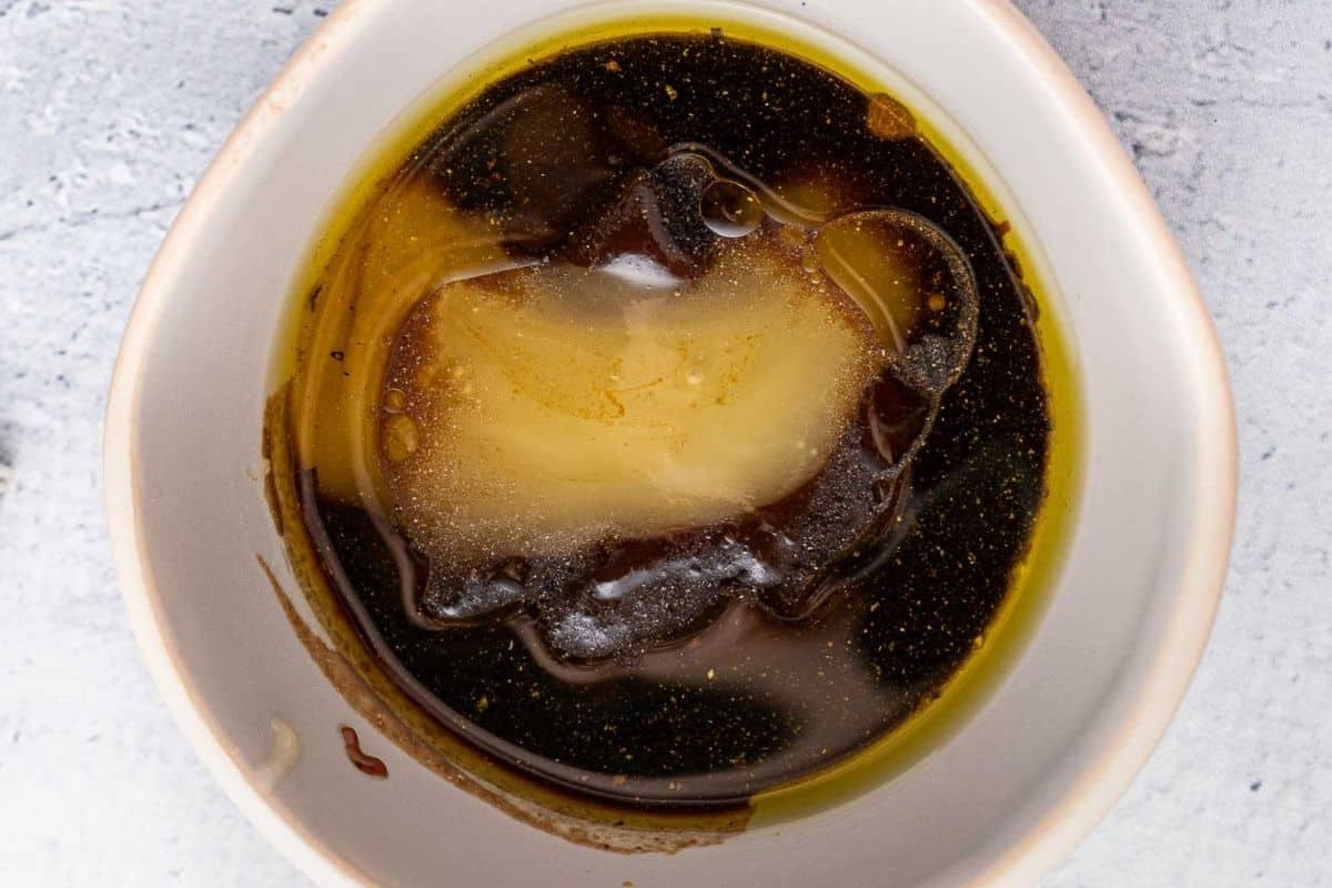 A close-up of a white bowl containing a mixture of soy sauce, teriyaki sauce, honey, and oil on a light-colored surface.