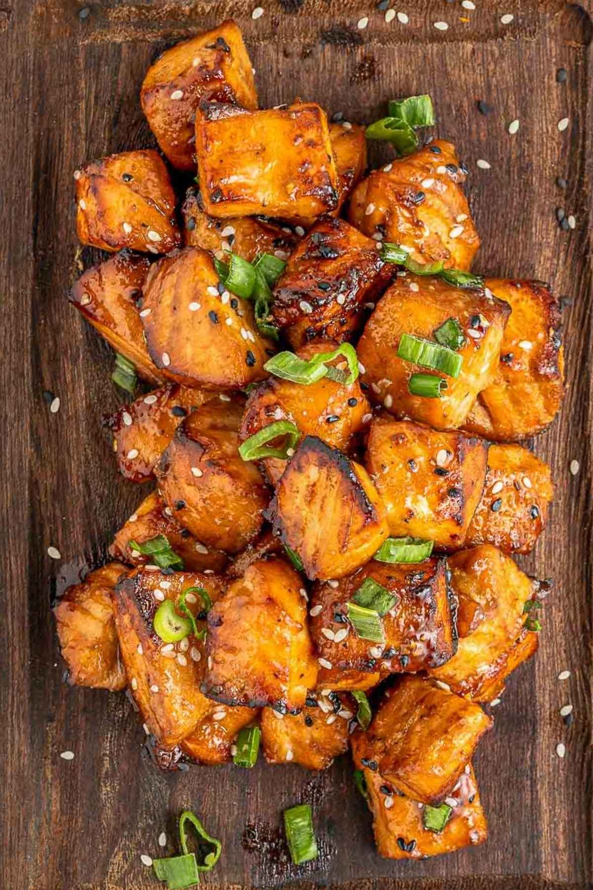 Crispy tofu cubes glazed with sauce, garnished with chopped green onions and sesame seeds, served on a wooden board.
