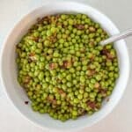 A white bowl filled with cooked green peas mixed with sautéed onions, with a spoon inside the bowl.