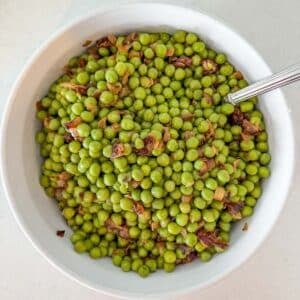 A white bowl filled with cooked green peas mixed with sautéed onions, with a spoon inside the bowl.