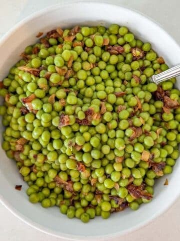 A white bowl filled with cooked green peas mixed with sautéed onions, with a spoon inside the bowl.