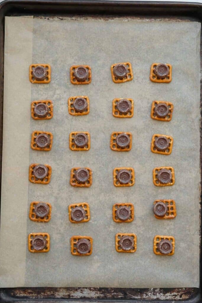 A baking sheet lined with parchment paper holds rows of square pretzels, each topped with a rolo.