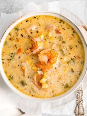 A bowl of creamy shrimp and corn chowder, garnished with whole shrimp, served with a spoon and fork on a white plate.