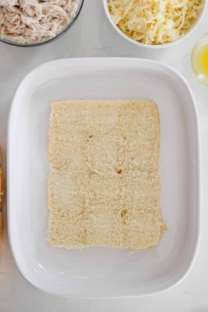 A layer of sliced Hawaiian bread roll bottoms arranged in a white rectangular baking dish.