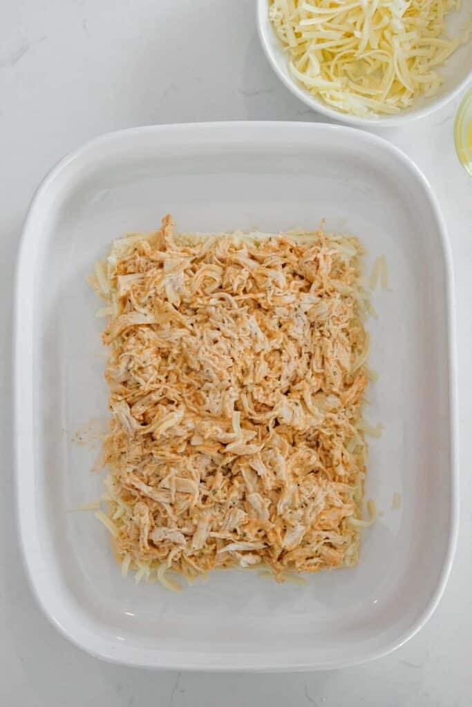 A white baking dish with a layer of shredded buffalo chicken over shredded cheese on the roll bottoms.