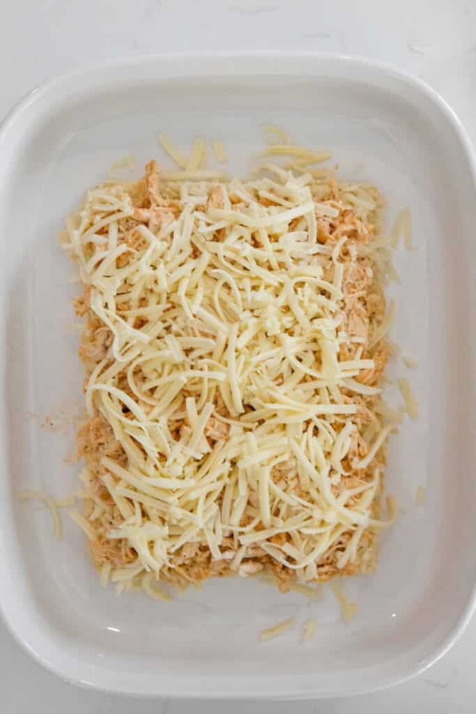 Rectangular white baking dish with a layer of shredded cheese on top of a buffalo chicken base on the bread rolls.