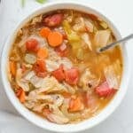 A white bowl of cabbage soup containing cabbage, carrots, celery, and tomatoes, with a spoon in the bowl.