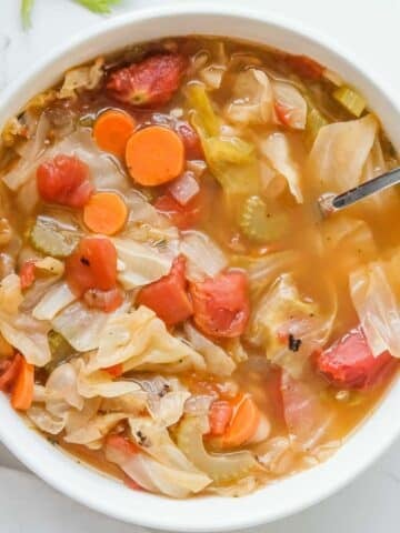 A white bowl of cabbage soup containing cabbage, carrots, celery, and tomatoes, with a spoon in the bowl.