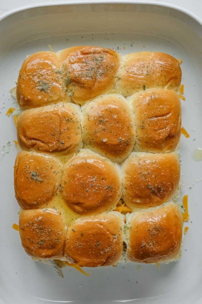 Tops of hawaiian rolls put back over the cheese and chicken mixture topped with herbs and melted butter, arranged neatly in a baking dish.